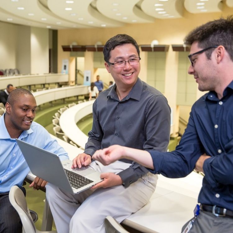 Three business professionals laugh together, looking at a laptop while they wait in the auditorium for conference to start.